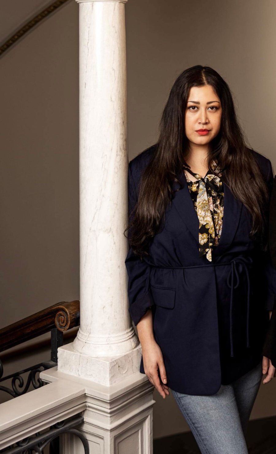 Louise Neo, grundare av Sistjejer, med svart hår och rött läppstift står lutat mot en pelare i svart kavaj.